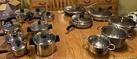Overview of multiple stainless steel pots and pans with lids arranged on a table including larger pots and smaller saucepans.