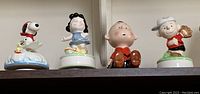 Four ceramic Snoopy and Peanuts character music figurines shown on a shelf with Snoopy on doghouse, Lucy, Charlie Brown, and another character