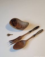 Displaying two wooden salad utensils with ornately designed metal handles, laid out beside a small wooden bowl with visible wood grain pattern and natural imperfection.