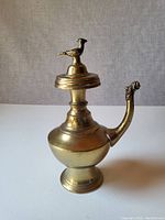 Front view of the antique brass tea pot with bird lid in place, showing detailed bird figure atop the lid and decorative spout.
