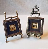 Two matching framed gold leaf artworks shown upright on different ornate stands on white background