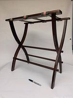 Front angled view of a dark wooden foldable suitcase stand with green and red straps on top, showing curved legs and crossbars for support.