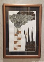 Frontal photo of framed abstract artwork showing tall column with hands and cloud shapes, and three black spires on the right.