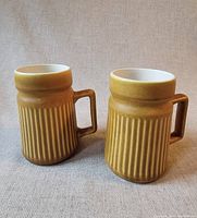Two large yellow ribbed ceramic mugs with square handles shown side by side on neutral fabric background.
