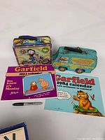 Overview of nostalgia lot showing Scooby Doo Mystery Machine tin, Betty Boop tin, two Garfield calendars from 1983 and 1984, and a black Sharpie marker.