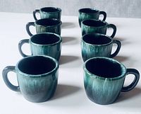 Front view of 8 blue-green glazed Blue Mountain Pottery mugs arranged in two rows.