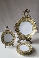 Photo showing one small side plate and one large dinner plate displayed on decorative metal stands, highlighting the handpainted gold and black fleur-de-lis border pattern on white porcelain with octagonal shape.