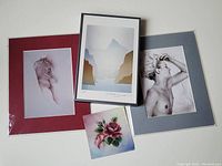 Photo showing all four art pieces including the framed 'Inlet' print, two matted prints with female nude figures, and the small square rose painting, laid flat on a surface.