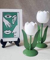 Photo showing two green and white tulip-shaped candle holders alongside the lithograph on a black easel