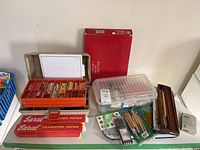 Top view showing a selection of pastel sticks, transfer paper boxes, plastic bins holding calligraphy brushes, drawing implements, and the Paterson Contact Proof Printer box in red behind the arrangement.