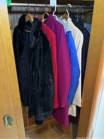 Seven assorted ladies coats and jackets hanging in a wooden closet rod including a black reversible faux fur coat, red, fuchsia wool coats, blue padded jacket, beige and navy coats.