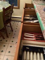 Open kitchen drawers showing multiple sets of silverplate flatware including knives with white handles, other cutlery and a cloth roll storage.