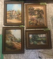 Four framed oil paintings arranged on floor, depicting cottages, trees, and water landscapes.