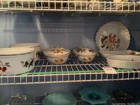 Shelf display of Royal Worcester ceramic serving bowls, lidded casseroles, and pie plate with floral and fruit designs, showing gold trim details.
