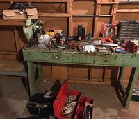 View of green wooden workbench with assorted tools including saws, clamps, gloves, flashlights, spotlight, and hardware strewn on top.