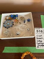 Top view of costume jewelry lot including brooches, earrings, ring, and beaded necklace. Includes note indicating Triad stamped brooch and vintage costume jewelry.