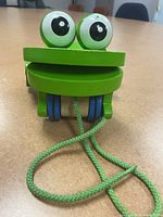 Front view of Melissa & Doug wooden Frolicking Frog pull toy, green with large eyes and blue wheels, showing pull string.