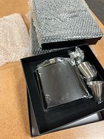 Top-down view of one black flask set packaged with a flask and two metal cups inside a black box, bubble wrap visible around the box.