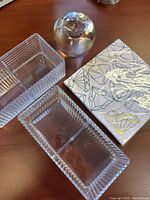 Three clear glass trays and a round glass apple paperweight on a wooden surface alongside a pack of diamond patterned napkins