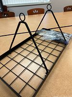 Full view of the black metal hanging rack showing the grid pattern and two triangular hooks with circular loops for hanging.