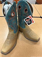 Full side and top view of the cowboy boots showing brown foot and teal blue shaft with orange and brown embroidery and circular pull holes, tags still attached indicating unworn condition.
