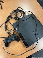 Sony PlayStation 3 console with wired DualShock 3 controller and cables on a tabletop.