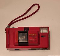 Front view of the red Safari Starshot 35mm film camera showing the lens, built-in flash, on/off switch, and wrist strap.