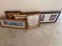 Five framed art pieces and plaques arranged leaning against a wall; various sizes and frame styles visible.