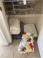 Closet shelf with 9 clear plastic stackable storage bins and pile of blankets and quilts on floor including leaf pattern quilt, seashell bed cover, white and dark gray blankets