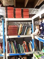 Three-tiered shelf holding multiple vintage books, predominantly red volumes on top with additional books on middle and lower shelves. Some dolls are perched on the shelf sides.