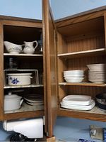 Cabinet with white plates, bowls, ceramic pitcher with floral pattern, Pfaltzgraff covered ovenware dish, and various kitchen tools