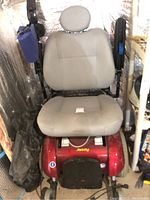 Full view of Jazzy electric wheelchair with beige padded seat and headrest, located indoors, surrounded by other items.