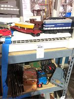 Photo showing Lionel model train layout on shelving with engine, caboose, cars, track and power transformer below.