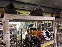 Metal shelving rack holding multiple power tools including belt sander, circular saws, electric drills, and router, stored in basement.