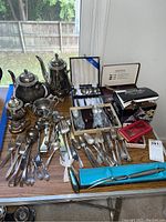 Wide view of entire lot showing teapots, flatware, buffet caddies, and boxes on table indoors near window.