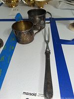 Photo of two vintage sterling silver cups and a sterling silver meat fork. The cups show tarnish; one cup has a handle, the other does not. The fork has two prongs and a textured handle.