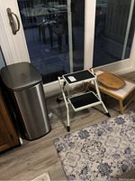 Photo of stainless steel trash can, two-step stool, wooden TV tray, and floor mat near glass door.