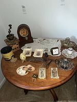 Full view of the round wooden table with all lot items including clock, coffee grinder, glasses, silver plate items, wall art, doily, perfume decanters