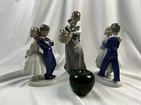 Front view showing the three porcelain figurines arranged with a green glass apple for size reference. Two Bing Grondahl young couple figurines in blue and white, and one Lladro young lady figure in blue and white with a basket of puppies.