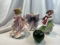Three Royal Doulton figurines shown with a green apple for scale. The figures are finely detailed depicting women in elegant period attire with color variations in dresses and distinct facial features.