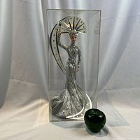 Front view of the doll in clear display case with green glass apple size reference