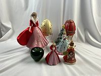 All six Christmas ornaments and figurines arranged together on white backdrop including lady figurines, musical eggs, Christmas tree, and Coca-Cola bell.