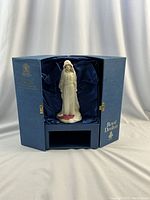 Open blue Royal Doulton box lined with navy satin holding white porcelain figurine of a veiled woman on a magenta-flocked base