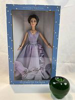Full view of doll in original blue box with white diamond pattern and green apple placed beside for size reference.