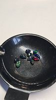 Photo shows multiple oval cut Ethiopian black opal gemstones in a black dish, showcasing their rainbow flash colors in green, blue, and red.