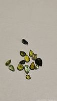 Top down photo of 14 small natural green tourmaline gemstones in varying shades and mixed cuts on a plain background.