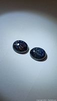 Pair of deep blue Pietersite gemstones shown on a white background under bright light with visible swirl patterns.