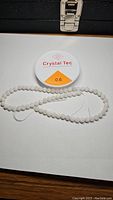 Natural white moonstone bead strand coiled alongside a spool of 0.6mm Crystal Tec clear stretchy thread on white surface.