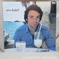 Front cover of Steve Hackett 'Cured' vinyl album featuring a photo of Steve Hackett sitting at a table by the sea with two glasses and the title 'Cured'.