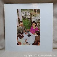 Front of the album cover showing four people sitting around a table with a black object, white border.